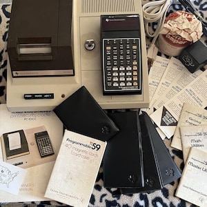 May include: A vintage Texas Instruments PC-100C print cradle and TI-59 programmable calculator set. Includes a beige thermal printer, black calculator, magnetic cards, multiple instruction manuals, paper rolls, and black protective carrying cases.