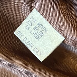 Puede incluir: Una etiqueta de cuidado rectangular blanca cosida sobre un forro de tela acolchada marr&oacute;n. El texto dice: 71% RAYON, 23% NYLON, 5% LYCRA, DRY CLEAN ONLY. La etiqueta est&aacute; orientada en diagonal sobre el fondo de tela tejida marr&oacute;n oscuro.
