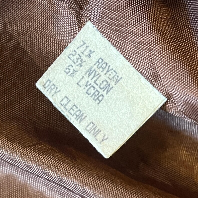 Puede incluir: Una etiqueta de cuidado rectangular blanca cosida sobre un forro de tela acolchada marr&oacute;n. El texto dice: 71% RAYON, 23% NYLON, 5% LYCRA, DRY CLEAN ONLY. La etiqueta est&aacute; orientada en diagonal sobre el fondo de tela tejida marr&oacute;n oscuro.
