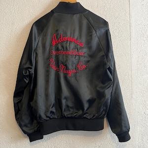 May include: Black satin bomber jacket with red embroidered text on the back. The text reads "Advance Overhead Door Van Nuys, Ca." The jacket has a black ribbed collar, cuffs, and hem. The jacket is a classic style.