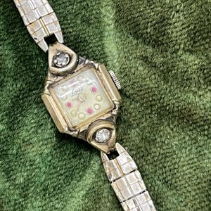 Puede incluir: Reloj plateado vintage con esfera rectangular y correa plateada. La esfera del reloj es de color crema con la palabra "Louis" en cursiva y pequeños detalles rojos y transparentes. El reloj tiene una piedra con forma de diamante a cada lado de la esfera.