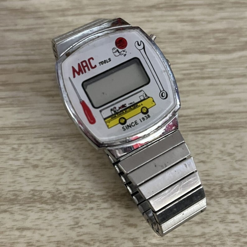 Op de afbeelding: Vintage MAC TOOLS digitale horloge met een zilveren band en witte wijzerplaat. De wijzerplaat toont het MAC TOOLS-logo, een schroevendraaier, een sleutel en een gele vrachtwagen met de tekst "SINCE 1938". Het horloge heeft een rechthoekig digitaal display.