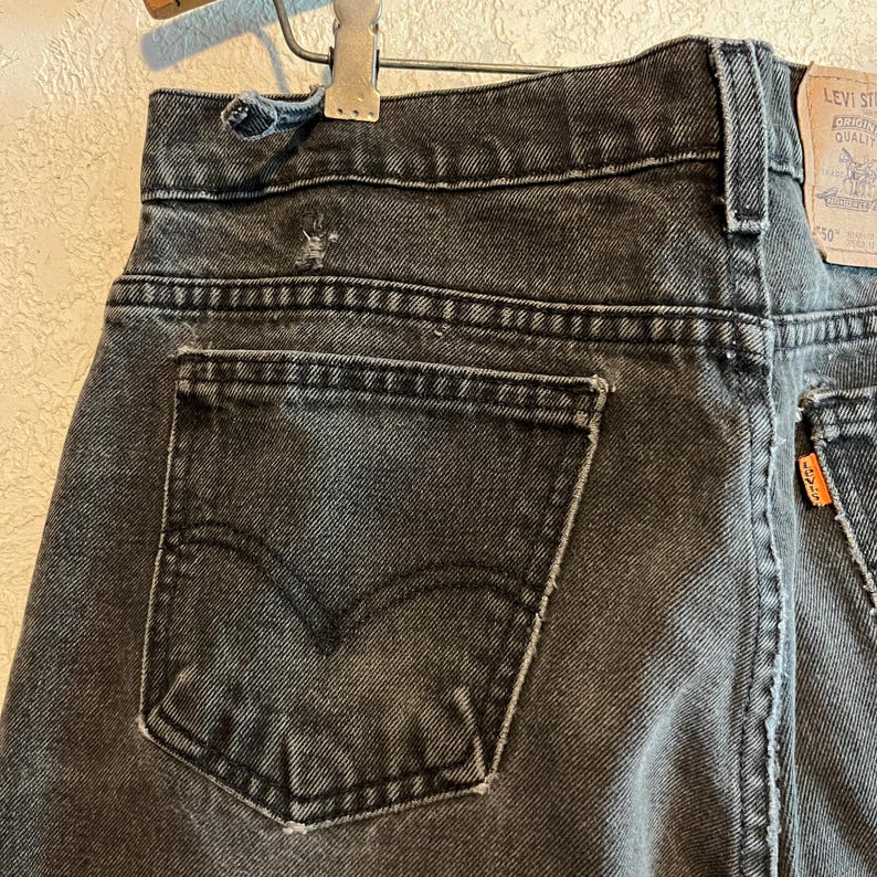 K&ouml;nnte beinhalten: Nahaufnahme der R&uuml;ckseite einer verblichenen schwarzen Jeans. Die Jeans hat ein klassisches F&uuml;nf-Taschen-Design mit einer Ges&auml;&szlig;tasche auf der linken Seite. Das Markenetikett von Levi's ist auf der rechten Seite sichtbar. Die Jeans ist abgenutzt, mit Gebrauchsspuren.