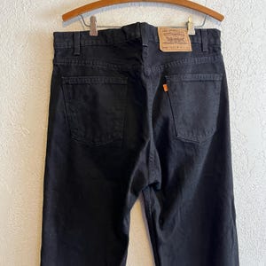 Puede incluir: Vaqueros negros de mezclilla con un diseño clásico de cinco bolsillos. Los vaqueros presentan un aspecto descolorido y una etiqueta naranja de Levi's en el bolsillo trasero. Los vaqueros están colgados en una percha de madera.