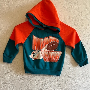 Puede incluir: Una sudadera con capucha de color azul verdoso y naranja con las palabras "Miami Dolphins" y un gráfico de fútbol impreso en la parte delantera.