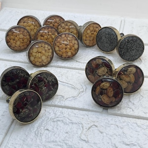 May include: A selection of cabinet knobs with diverse designs. Some knobs feature dried beans encased in clear resin, while others have a speckled grey stone appearance. The knobs have a silver-coloured metal base.