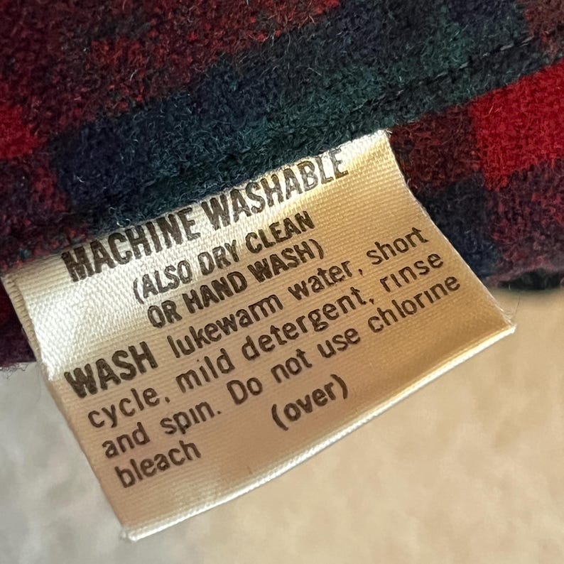 May include: A fabric care label attached to a red and dark green plaid garment. The label reads: MACHINE WASHABLE (ALSO DRY CLEAN OR HAND WASH) WASH lukewarm water, short cycle, mild detergent, rinse and spin. Do not use chlorine bleach (over).