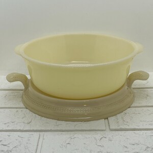 Puede incluir: Un cuenco de vidrio amarillo vintage con dos asas, sobre una base decorativa beige. La base tiene un diseño en relieve detallado. Esta pieza es probablemente una fuente para servir o una cazuela, perfecta para la decoración de cocina retro. Diámetro del cuenco: aprox. 20 cm.