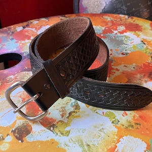 Puede incluir: Un cinturón de cuero marrón oscuro con un patrón en relieve de tejido de cesta que se repite a lo largo de la correa. El cinturón incluye una trabilla de cuero liso y una hebilla de metal plateado pulido con un solo perno, fijada con un remache.