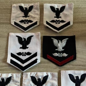 Könnte beinhalten: Eine Sammlung bestickter militärischer Rangabzeichen mit einem Adler über gekreuzten Ankern. Die Abzeichen sind weiß mit schwarzer Stickerei oder schwarz mit weißer und roter Stickerei und zeigen jeweils Rang-Chevron-Streifen.