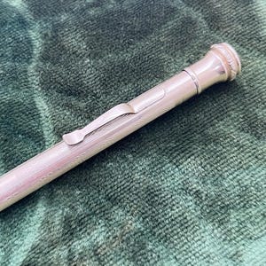 Könnte beinhalten: Ein silberfarbener Druckbleistift mit Clip und dekorativen Details. Der Stift liegt auf einer grünen, strukturierten Oberfläche. Das Design des Stifts umfasst einen Clip und einen strukturierten Körper.