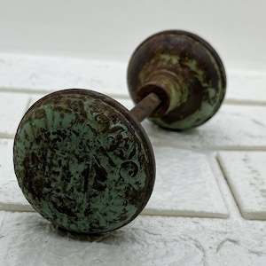 Peut inclure: Bouton de porte ancien avec une finition vieillie et patinée. Le bouton est de couleur bronze foncé avec des zones de patine verte. Le design présente une forme arrondie avec des détails décoratifs. Le bouton est fixé à une tige métallique.