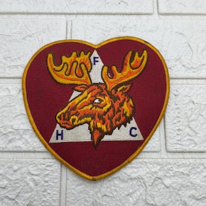 May include: A heart-shaped patch with a maroon background and a gold border. The patch features a detailed illustration of a moose with orange and yellow fur. The letters "F", "H", and "C" are visible.
