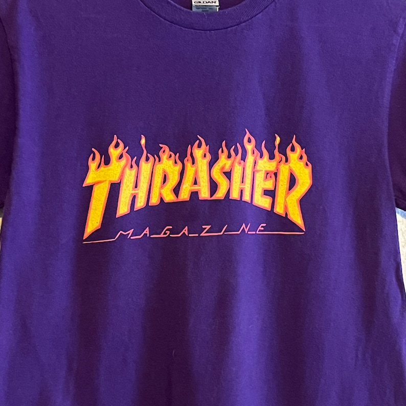 May include: Purple t-shirt featuring a yellow and orange "Thrasher Magazine" logo with flames.