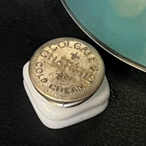 以下が含まれることがあります： 白いセラミックベースと金属製の蓋が付いた、ビンテージのコルゲートコールドクリームジャー。蓋にはブランド名と製品の詳細がエンボス加工されています。このジャーは、化粧品やスキンケア製品を示唆する、クラシックでアンティークな美学を持っています。