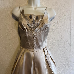 May include: A champagne-colored satin dress with a fitted bodice and a full skirt. The bodice features a beaded embellishment and a row of small buttons down the front. The dress has thin spaghetti straps and a V-neckline.