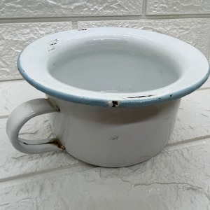 May include: White enamel chamber pot with a rounded rim and a handle. The rim is trimmed with a light blue accent. The pot shows signs of wear and tear, with some areas of chipped enamel.