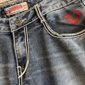 Pu&ograve; includere: Primo piano di jeans blu in denim con cuciture bianche e un logo a ferro di cavallo rosso sulla tasca. I jeans presentano una chiusura con bottoni e un'etichetta che recita "TRUE RELIGION BRAND JEANS WORLD TOUR FASHION FOR THE SENSES."