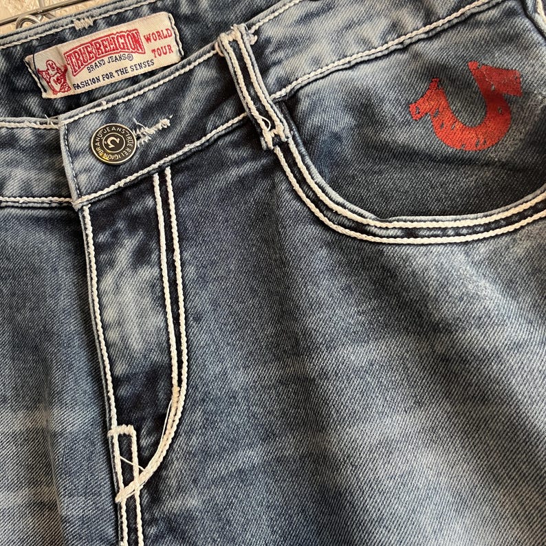 Pu&ograve; includere: Primo piano di jeans blu in denim con cuciture bianche e un logo a ferro di cavallo rosso sulla tasca. I jeans presentano una chiusura con bottoni e un'etichetta che recita "TRUE RELIGION BRAND JEANS WORLD TOUR FASHION FOR THE SENSES."