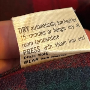 May include: A cream-colored fabric care label with black text that reads: DRY automatically, low heat for 15 minutes or hanger dry at room temperature. PRESS with steam iron and press cloth. WEAR with pleasure. The label is attached to a plaid fabric.