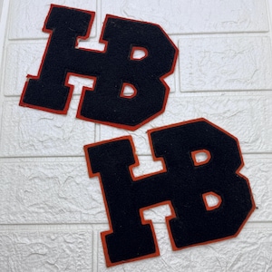 May include: Two varsity-style letter patches featuring the letters HB in a bold, block font. Each patch has a dark navy blue felt center with a vibrant orange border outlining the shape of the letters, creating a high-contrast collegiate appearance.