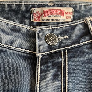 Pu&ograve; includere: Primo piano di jeans blu con cuciture bianche. I jeans presentano una chiusura con bottone e un'etichetta del marchio con la scritta "TRUE RELIGION" e "FASHION FOR THE SENSES". I jeans sono uno stile classico.