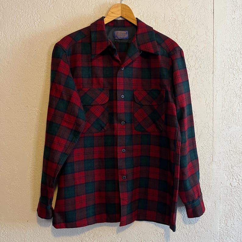 May include: A long-sleeve button-up shirt featuring a dark red and forest green plaid pattern. The shirt includes a pointed collar, two chest pockets with flaps, and a central row of dark buttons. A Pendleton brand label is visible at the neck.