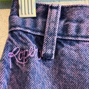 May include: Close-up of a pair of denim jeans in a blue and purple wash. The jeans feature a belt loop and embroidered lettering in a cursive font. The fabric has a textured appearance.