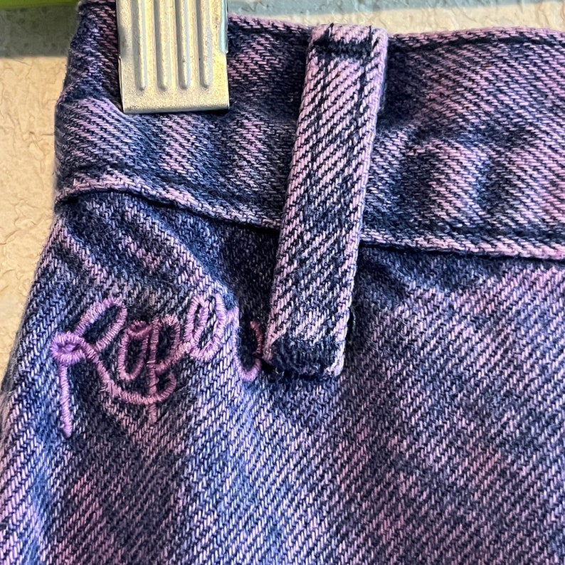 May include: Close-up of a pair of denim jeans in a blue and purple wash. The jeans feature a belt loop and embroidered lettering in a cursive font. The fabric has a textured appearance.