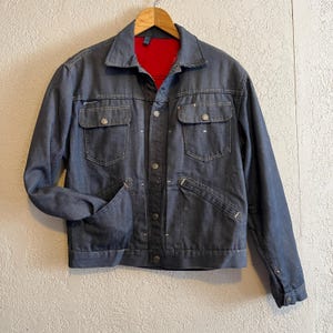 May include: A denim jacket with a dark blue exterior and a red interior lining. The jacket features two chest pockets, two side pockets, and a button-up front. It is hanging on a wooden hanger.
