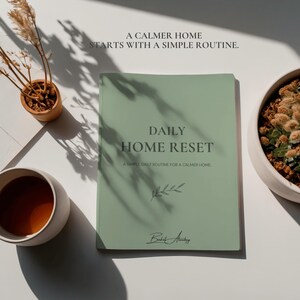 Puede incluir: Un libro de color verde salvia titulado DAILY HOME RESET A SIMPLE DAILY ROUTINE FOR A CALMER HOME con una pequeña ilustración de una hoja y una firma en la parte inferior. El texto A CALMER HOME STARTS WITH A SIMPLE ROUTINE aparece arriba.