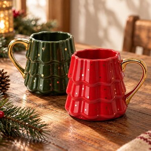 Puede incluir: Dos tazas de cerámica con forma de árboles de Navidad escalonados, una de color verde oscuro y otra de color rojo brillante. Ambas tazas presentan un acabado brillante, asas doradas y pequeños puntos dorados dispersos por la superficie exterior.