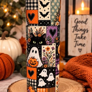 May include: A cylindrical tumbler featuring a patchwork design with Halloween motifs including black cats, white ghosts, orange pumpkins, bats, and floral patterns. The print incorporates black, orange, purple, and cream colors in a grid-like arrangement.