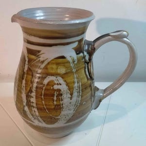 May include: A ceramic pitcher featuring a wide, flared rim and a curved handle. The exterior displays a hand-painted pattern of swirling white brushstrokes layered over a warm, earthy brown glaze, creating a textured, multi-tonal appearance.