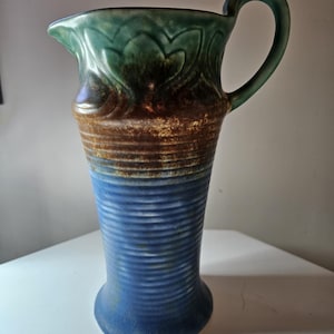 May include: A ceramic pitcher featuring a flared rim and a curved handle. The upper section displays a green glaze with a moulded leaf pattern, transitioning into a brown band, and ending in a ribbed blue base with a matte, textured finish.