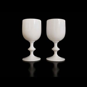 May include: Two matching white opaque glass goblets with a smooth, rounded bowl, a slender stem featuring a central decorative ring, and a wide, circular base. The glasses have a glossy finish and are positioned side by side against a black background.