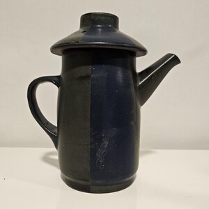 May include: A ceramic teapot featuring a two-tone design with one side in matte charcoal grey and the other in a deep, glossy navy blue. The teapot includes a matching lid with a central knob, a curved handle on the left, and a tapered spout.