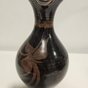 May include: A dark brown ceramic pitcher with a glossy finish and a flared, spout-like rim. The body of the pitcher features a lighter brown, stylised floral or leaf-like pattern etched or painted onto the surface, contrasting with the dark glaze.