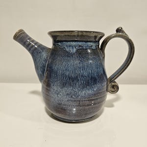 May include: A ceramic pitcher featuring a rounded body and a wide rim, finished in a variegated glaze of deep navy blue, slate grey, and lighter blue streaks. It includes a tapered spout on the left and a curved handle with a curled base detail.