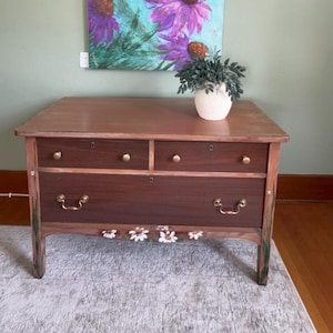 Op de afbeelding: Een houten ladekast met een donkerbruine afwerking, voorzien van twee kleine bovenlades en één grote onderlade. Het meubel heeft messing beslag, waaronder ronde knoppen en twee handgrepen. Een handgeschilderd bloemmotief met witte bloemen siert de onderste rand.