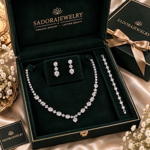May include: A jewellery set featuring a silver-toned necklace, a matching bracelet, and two stud earrings, all adorned with clear round stones. The set is presented in a dark green velvet-lined box with the text SADORAJEWELRY printed in gold.