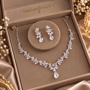 May include: A silver-toned jewellery set featuring a necklace and matching drop earrings, each adorned with clear, marquise-cut and pear-shaped crystals in a leaf-like arrangement. The set is presented in a beige box labelled SadoraJewellery.