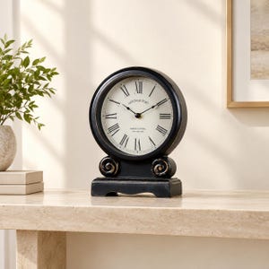 May include: A black mantel clock with a round face, cream-colored dial, and Roman numerals. The clock features the text "HERITAGE PORT" and "GRAND CENTRAL NEW YORK 1887" on the face, with a sturdy base and two decorative scroll accents.