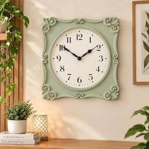 May include: A square wall clock with a sage green frame featuring decorative scrollwork at each corner. The clock face is off-white with black Arabic numerals from one to twelve and black hour and minute hands centered on the dial.