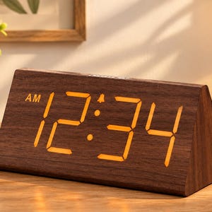 May include: A triangular digital alarm clock with a dark brown wood grain finish. The front face displays the time 12:34 in bright orange LED numbers, with the letters AM visible in the upper left corner of the display screen.