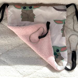 Peut inclure: Un hamac triangulaire pour animal avec une face supérieure en tissu blanc ornée d'un motif répétitif de personnage vert et une face inférieure en polaire rose douce. Il comporte des sangles élastiques noires et des anneaux métalliques argentés.
