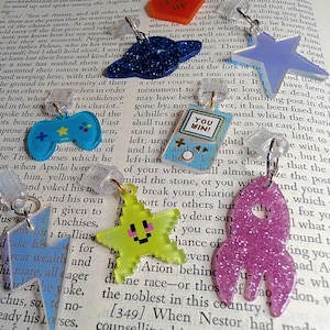 May include: A collection of eight acrylic charms featuring various shapes including a red heart with the text LEVEL UP, a blue glitter planet, a light blue star, a blue game controller, a handheld console with the text YOU WIN, a lightning bolt, a yellow pixelated star, and a pink glitter rocket.