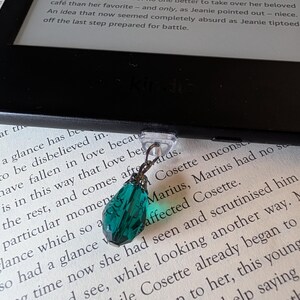May include: A dark teal, faceted, teardrop-shaped crystal charm attached to a clear plastic clip. The charm features a metallic cap and is suspended from the bottom edge of a black e-reader device, which rests on a page of printed text.