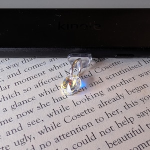 May include: A clear, faceted crystal charm attached to a small silver-toned metal ring, positioned against the bottom edge of a black Kindle e-reader device which rests on a page of printed text from a book featuring the names Marius and Cosette.