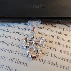 May include: A silver-toned metal dragon charm attached to a clear plastic clip. The dragon is depicted in a coiled position with visible wings, a long tail, and detailed scales. The charm is positioned against a page of printed text.